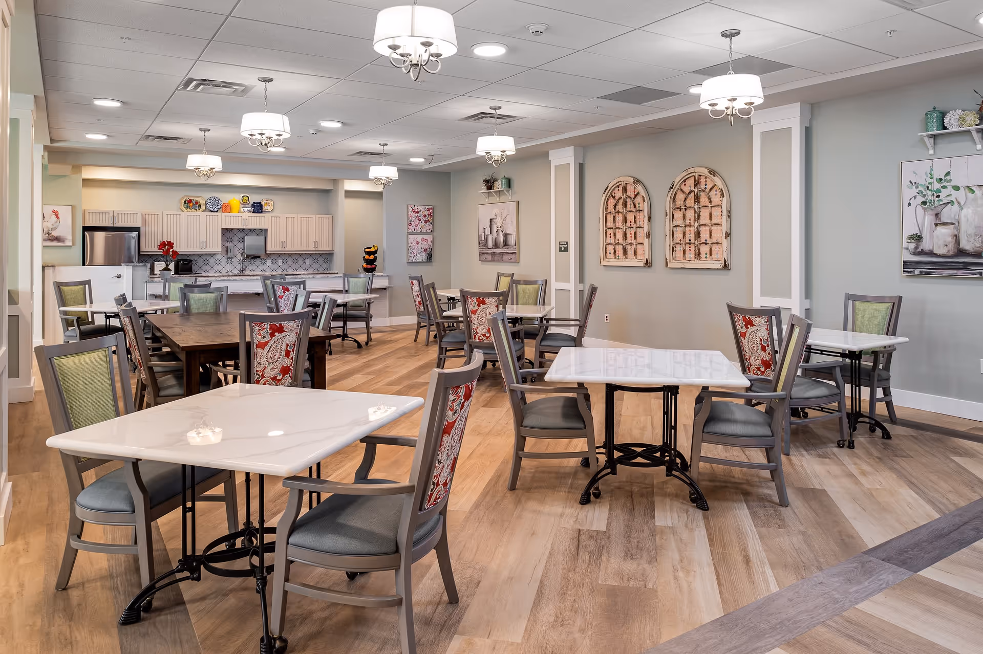 A dining room area in a senior living facility with multiple tables and chairs arranged neatly. The room features light-colored walls with decorative artwork, wooden flooring, and ceiling lights. In the background, there is a kitchenette with cabinets, a refrigerator, and a countertop with decorative items.
