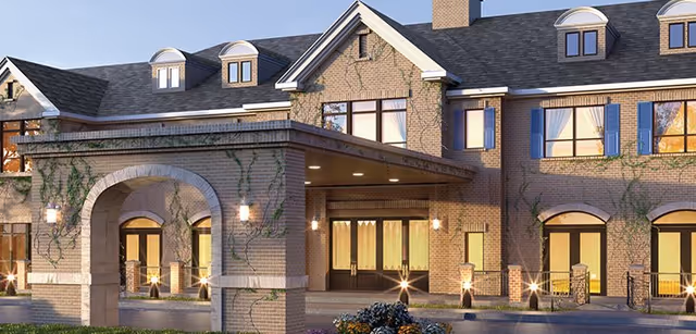 Exterior view of a senior living facility building with brick walls, multiple windows, and a covered entrance with arches and outdoor lighting. Ivy plants grow on the walls, and there are some flowers near the entrance.