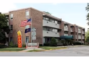 Front exterior of a three-story brick retirement community building with flagpoles and a sidewalk sign.