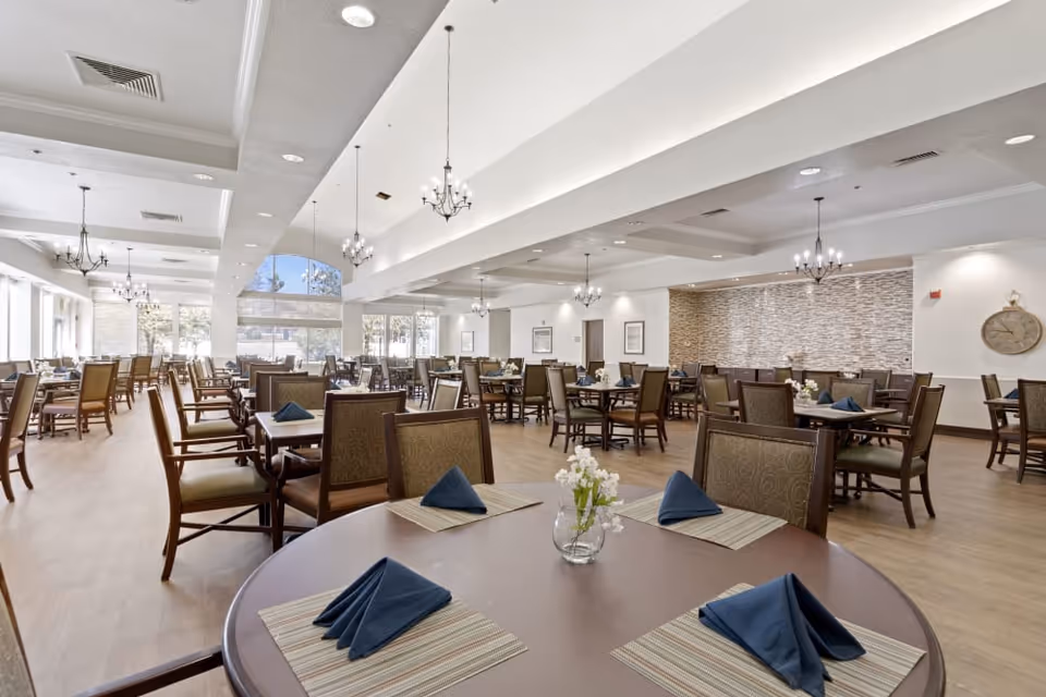 A spacious and well-lit dining room with multiple wooden tables and chairs arranged neatly. Each table is set with placemats, folded navy blue napkins, and small flower vases. The room features large windows allowing natural light to fill the space, chandeliers hanging from the ceiling, and a decorative clock on the wall.