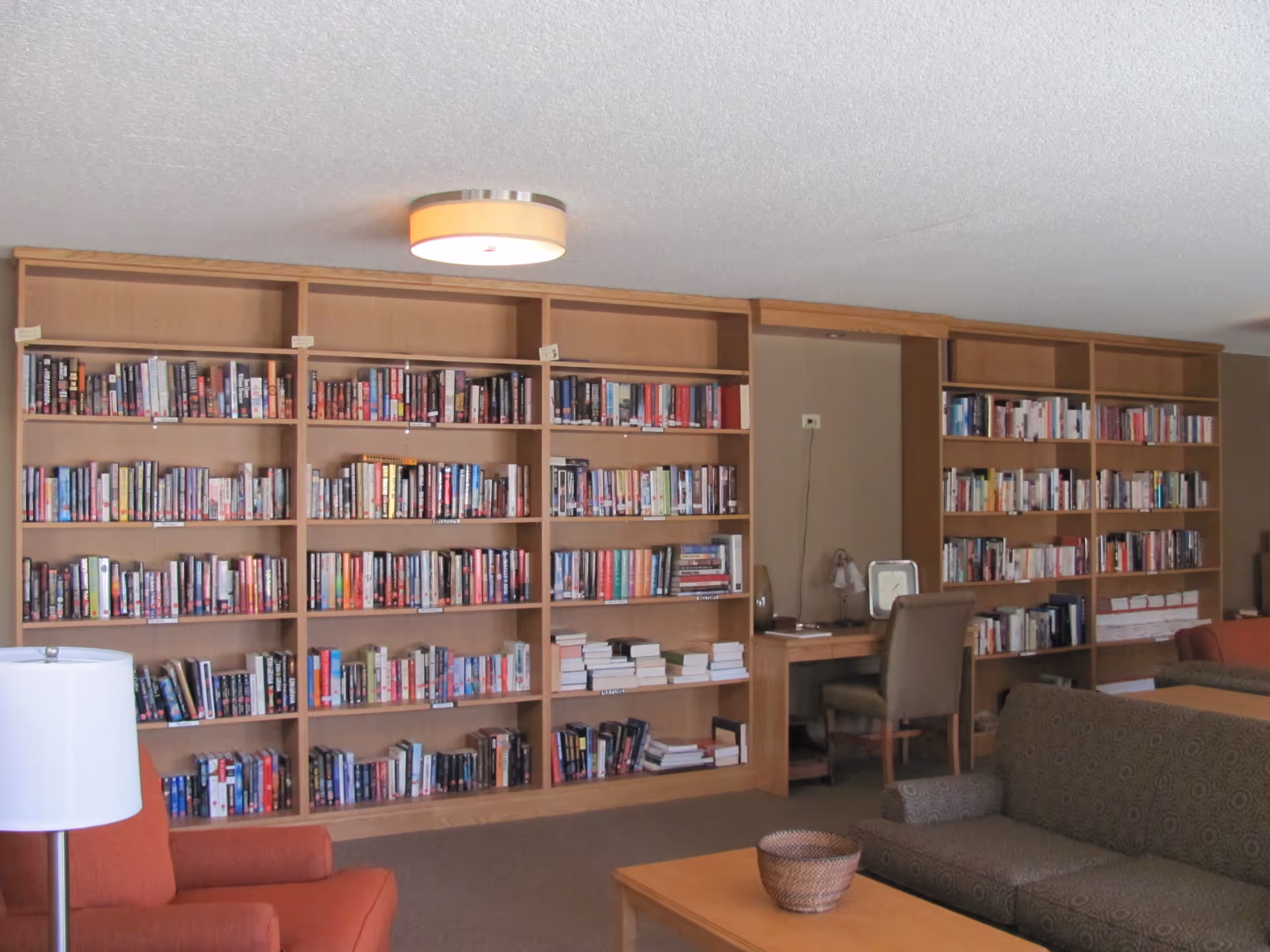 A cozy library room with large wooden bookshelves filled with books along the back wall. There is a wooden desk with a chair and a lamp in the middle section of the shelves. In the foreground, there is a brown patterned sofa, an orange armchair, a wooden coffee table with a woven basket, and a white floor lamp. The ceiling has a round light fixture.
