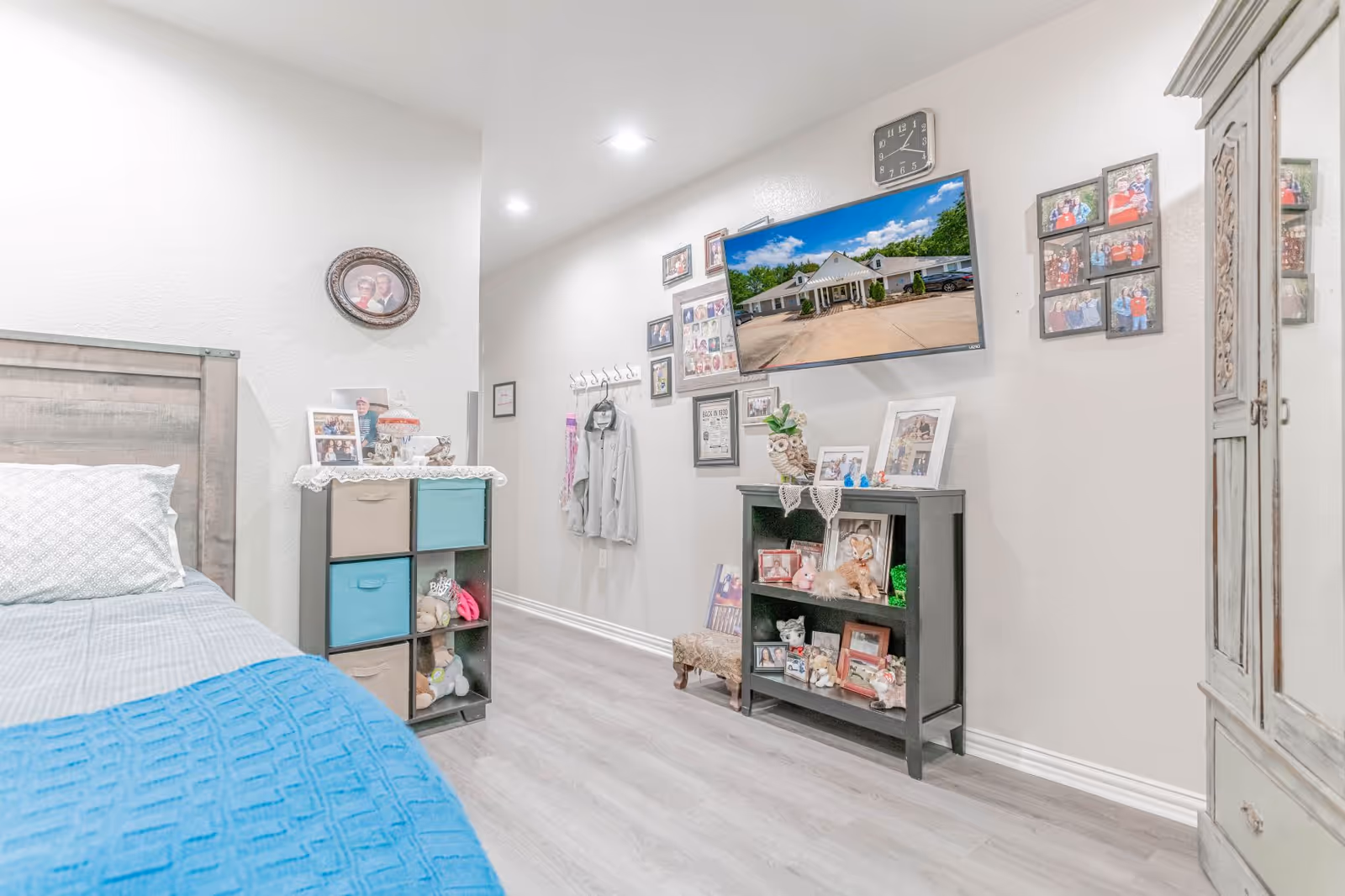 Bright furnished bedroom with a bed, cube storage, wall-mounted TV and photo-filled shelves along a light-colored wall.