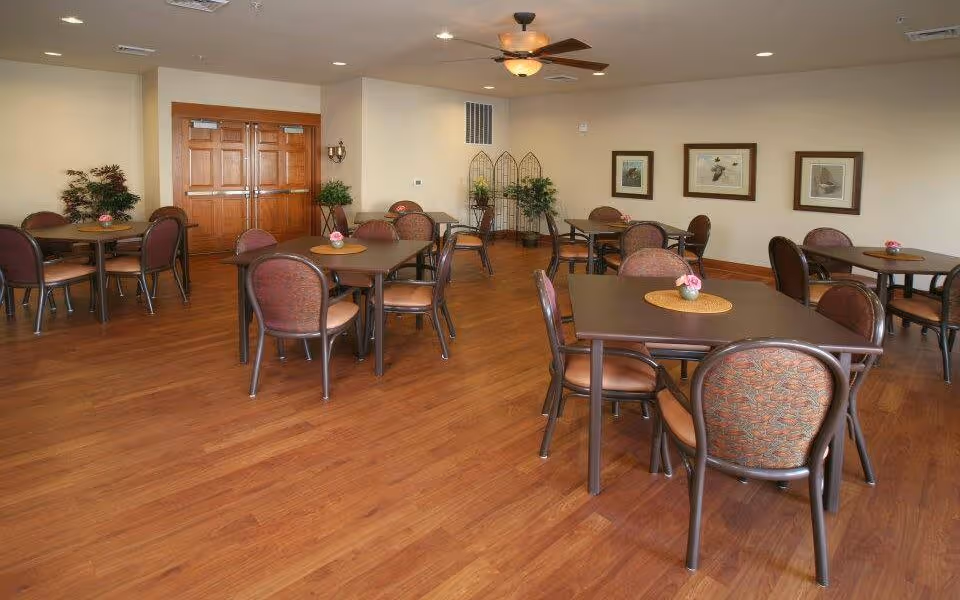 A spacious dining room with several square tables, each surrounded by four chairs with patterned upholstery. The room has wooden flooring, beige walls, and ceiling lights with a ceiling fan. There are framed pictures on the wall and some potted plants in the corner near double wooden doors.