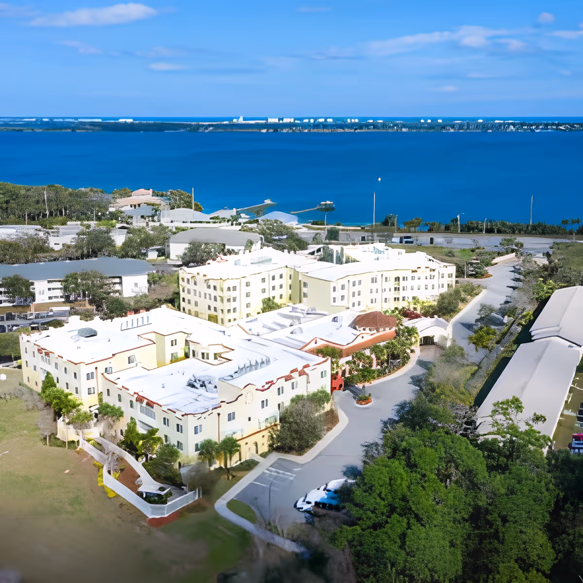 Aerial view of a senior living facility complex near a large body of water with multiple buildings surrounded by trees and greenery under a blue sky.
