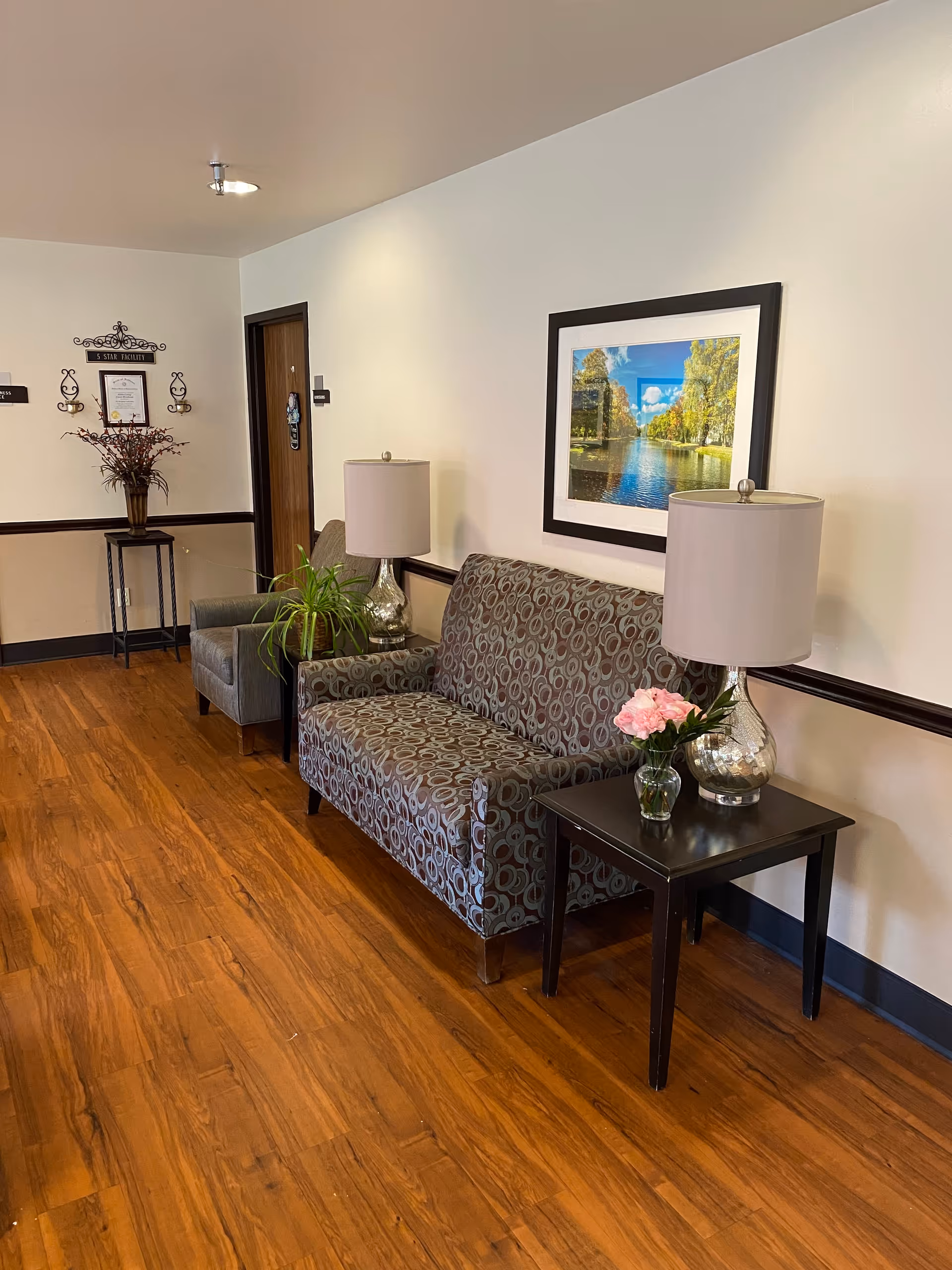A cozy seating area in a hallway with a patterned loveseat, a gray armchair, two table lamps with beige shades, a small side table holding a vase with pink flowers, and a framed landscape photograph on the wall. The floor is wooden, and there is a small table with a plant and decorative items near a door.