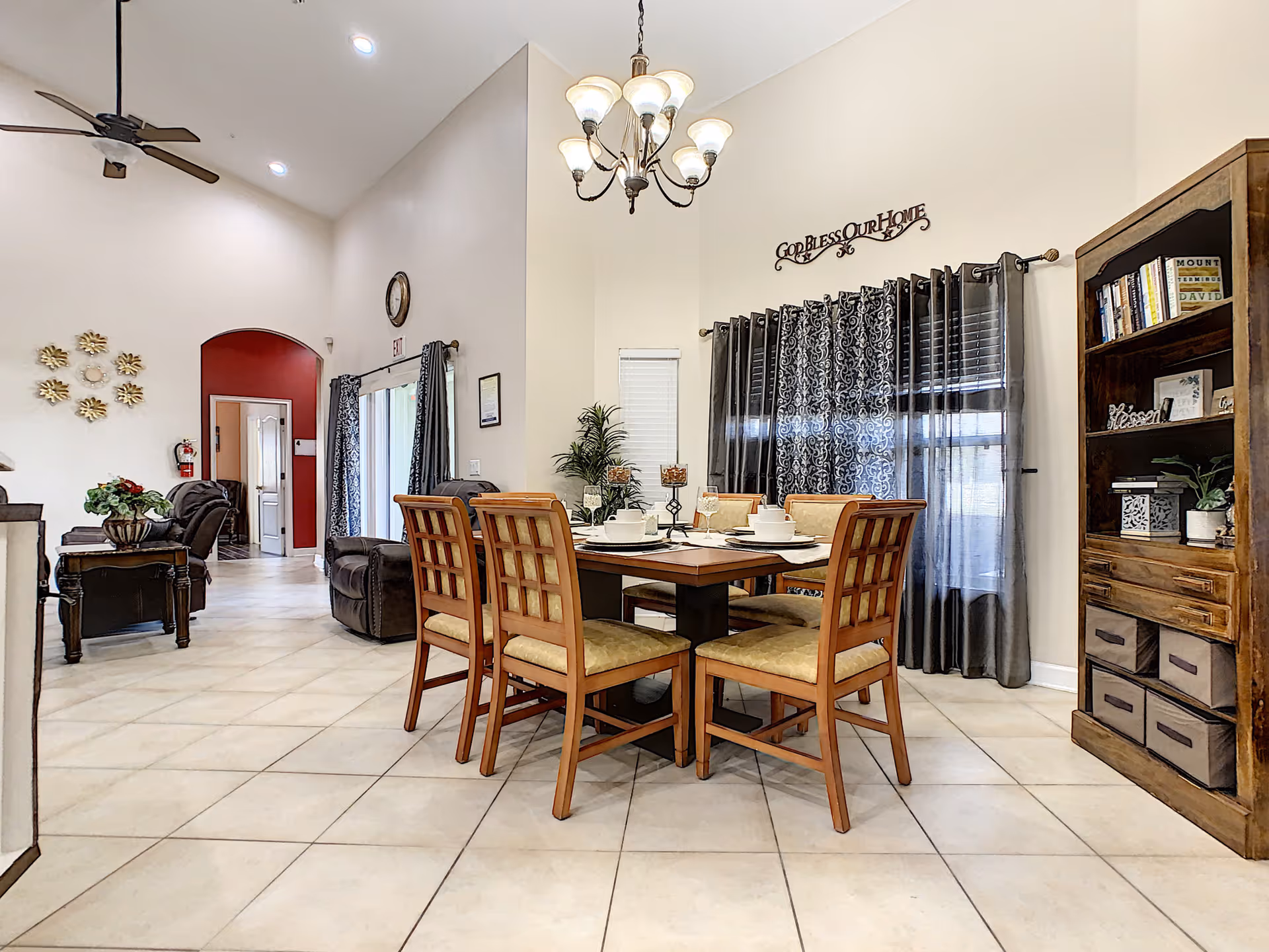 Interior view of a senior living facility dining area with a wooden dining table set for four, surrounded by cushioned chairs. The room has high ceilings with a chandelier, patterned curtains on the windows, a wooden bookshelf filled with books and decorative items, and a living area with recliner chairs visible in the background. The walls are light-colored with a decorative wall sign that reads 'God Bless Our Home'.