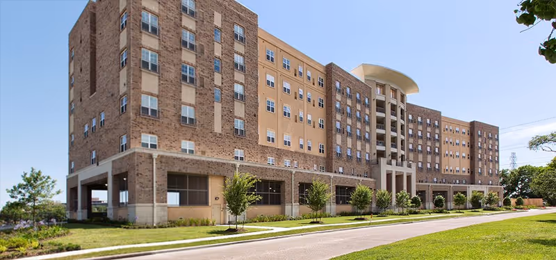 Large brick senior living building with manicured lawn beneath a clear blue sky.