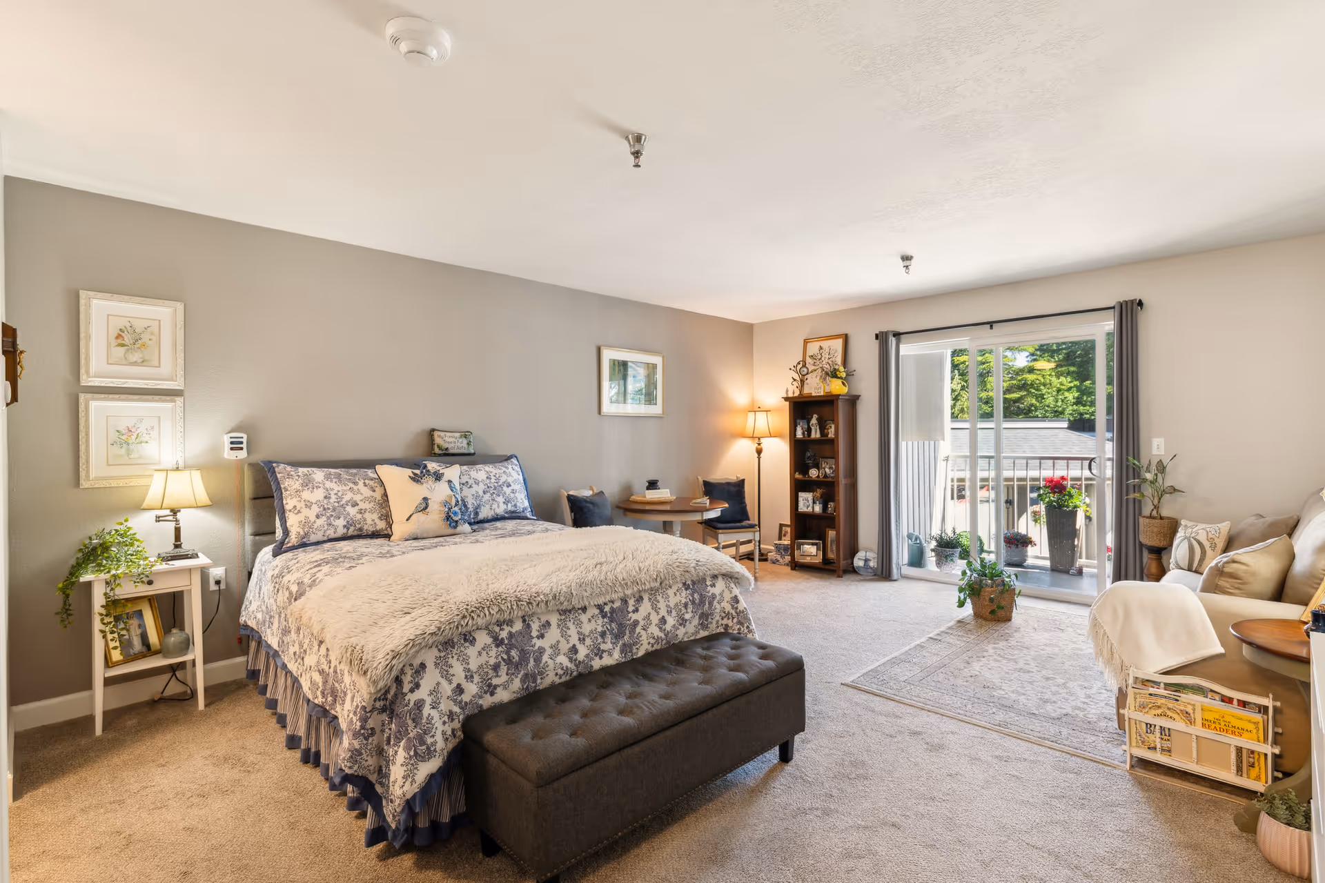 A spacious and well-lit bedroom in an assisted living facility featuring a large bed with floral bedding and a plush throw, a small side table with a lamp and plant, a cushioned bench at the foot of the bed, a small round table with two chairs in the corner, a tall bookshelf with decorative items, a sliding glass door leading to a balcony with potted plants, and a cozy seating area with a couch and side table.
