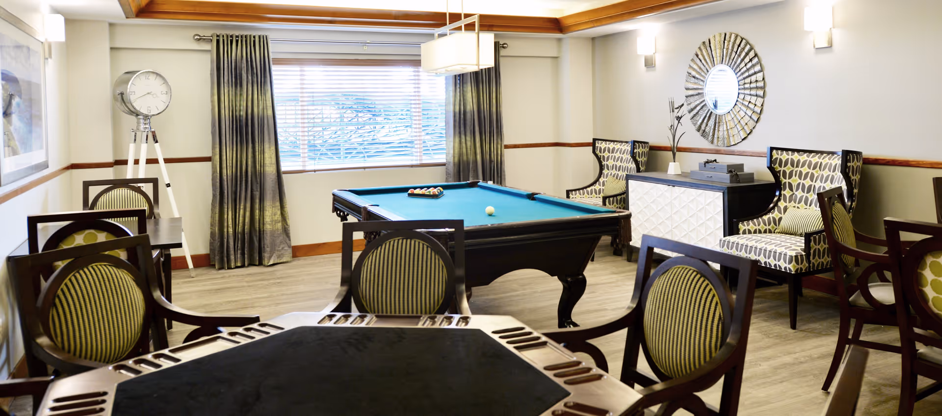 A well-lit recreational room with a pool table in the center, surrounded by several chairs and tables. The room features a large window with curtains, a tripod floor clock, patterned armchairs, a decorative mirror on the wall, and a sideboard with decorative items.