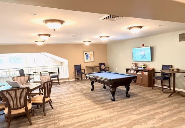A spacious recreational room with a pool table in the center, several chairs and tables arranged around the room, a wall-mounted TV displaying a game, and shelves with board games. The room has light-colored walls and ceiling lights, with a large window providing natural light.