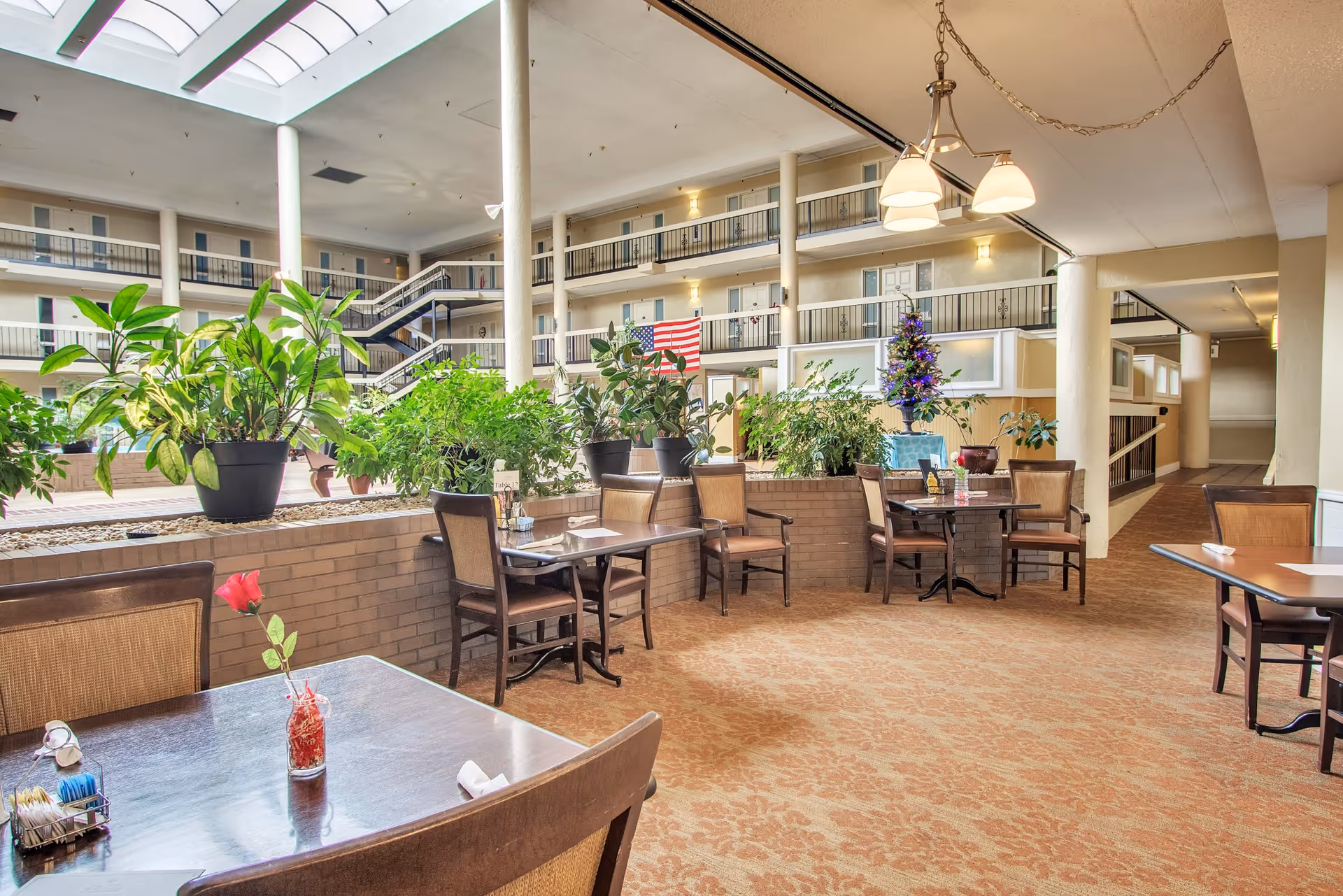 Indoor atrium dining area with tables and chairs, large potted plants, and multi-story balcony walkways.