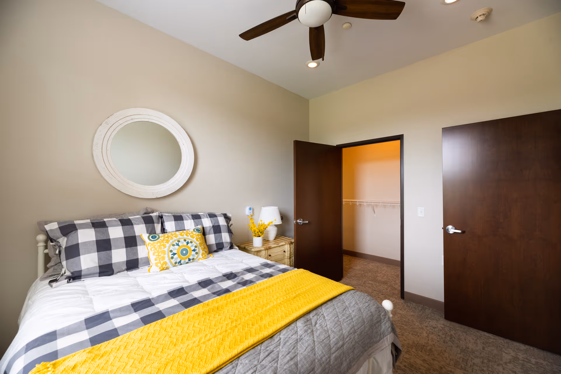 Bedroom with a bed dressed in gray-and-white checkered linens and a yellow throw, a round mirror above the headboard, a nightstand, ceiling fan, and an open closet with dark wood doors.