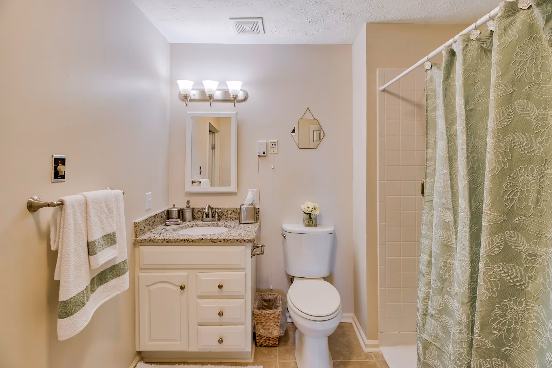 Well-lit bathroom with a vanity and sink, mirror, toilet, and a shower with a green patterned curtain.