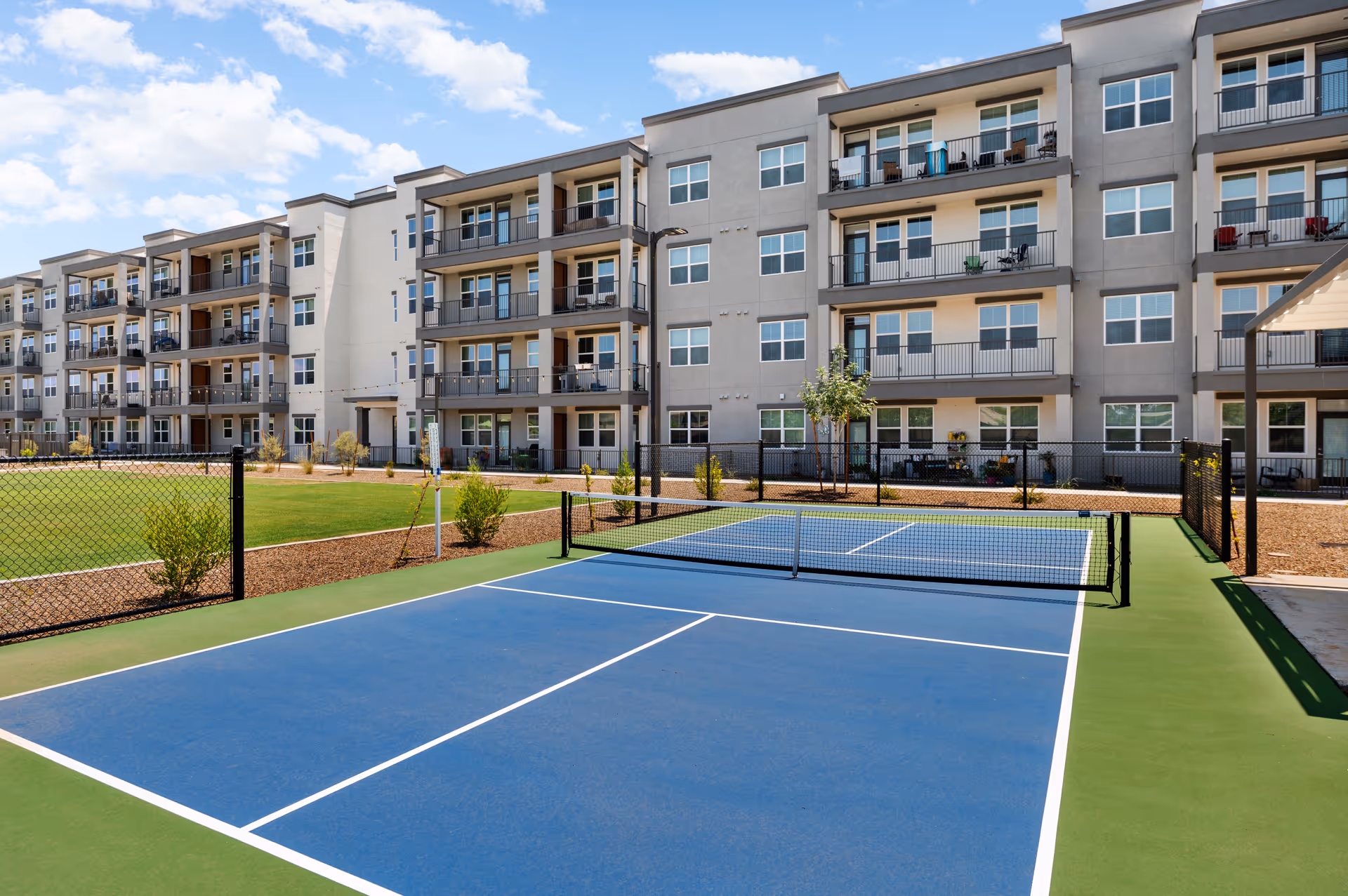 A blue tennis/pickleball court with a net in front of a four-story apartment building with balconies under a partly cloudy sky.