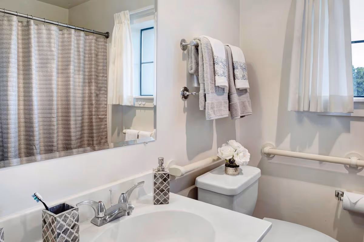 A clean and bright bathroom featuring a white sink with a silver faucet, a toothbrush holder, and a soap dispenser on the countertop. Above the sink is a large mirror reflecting a window with white curtains. To the right, there is a toilet with a small vase of white flowers on the tank. Two towel racks hold neatly folded towels, and grab bars are installed on the walls near the toilet and window for safety.