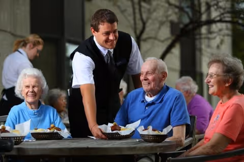 A young male server in a black vest and white shirt serves food baskets to three elderly people sitting around a table outdoors, all smiling and enjoying the moment.