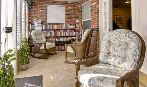 A cozy indoor seating area with three cushioned wicker chairs arranged near a bookshelf filled with books against a brick wall. There is a window with blinds above the bookshelf and a potted plant on the tiled floor near a glass door with vertical blinds.
