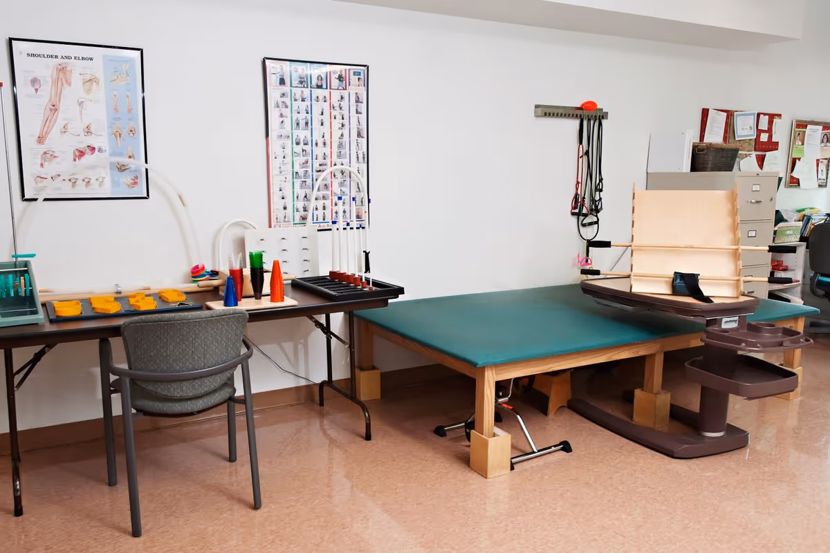 A therapy/rehab room with a treatment table, exercise equipment, a chair and a table holding therapy tools and instructional posters on the wall.