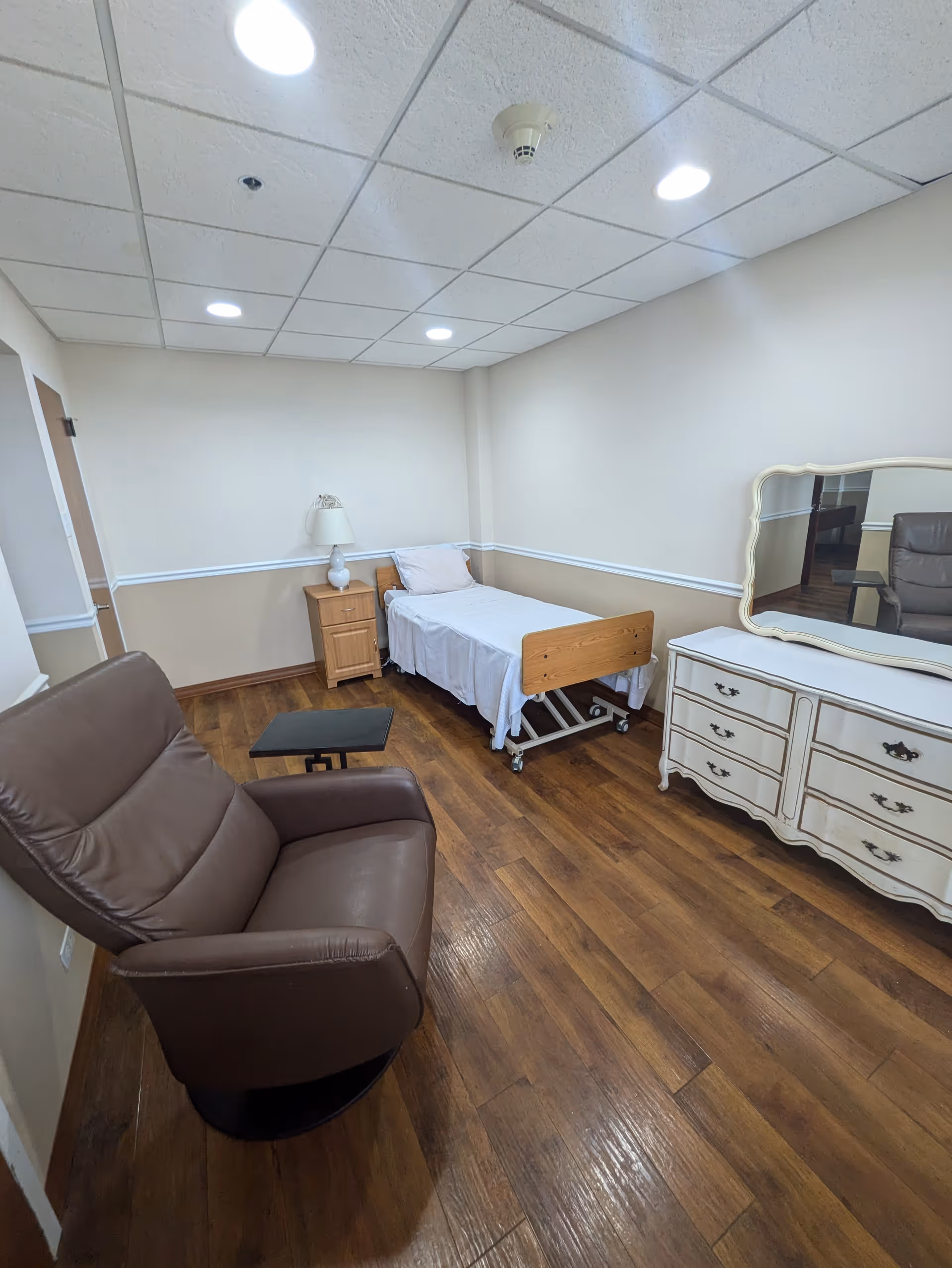 A small, simple bedroom with a single hospital-style bed with white sheets and a pillow, a wooden nightstand with a lamp, a brown leather recliner chair with a small black side table, and a white dresser with a large mirror. The room has wood flooring and beige walls with white trim.