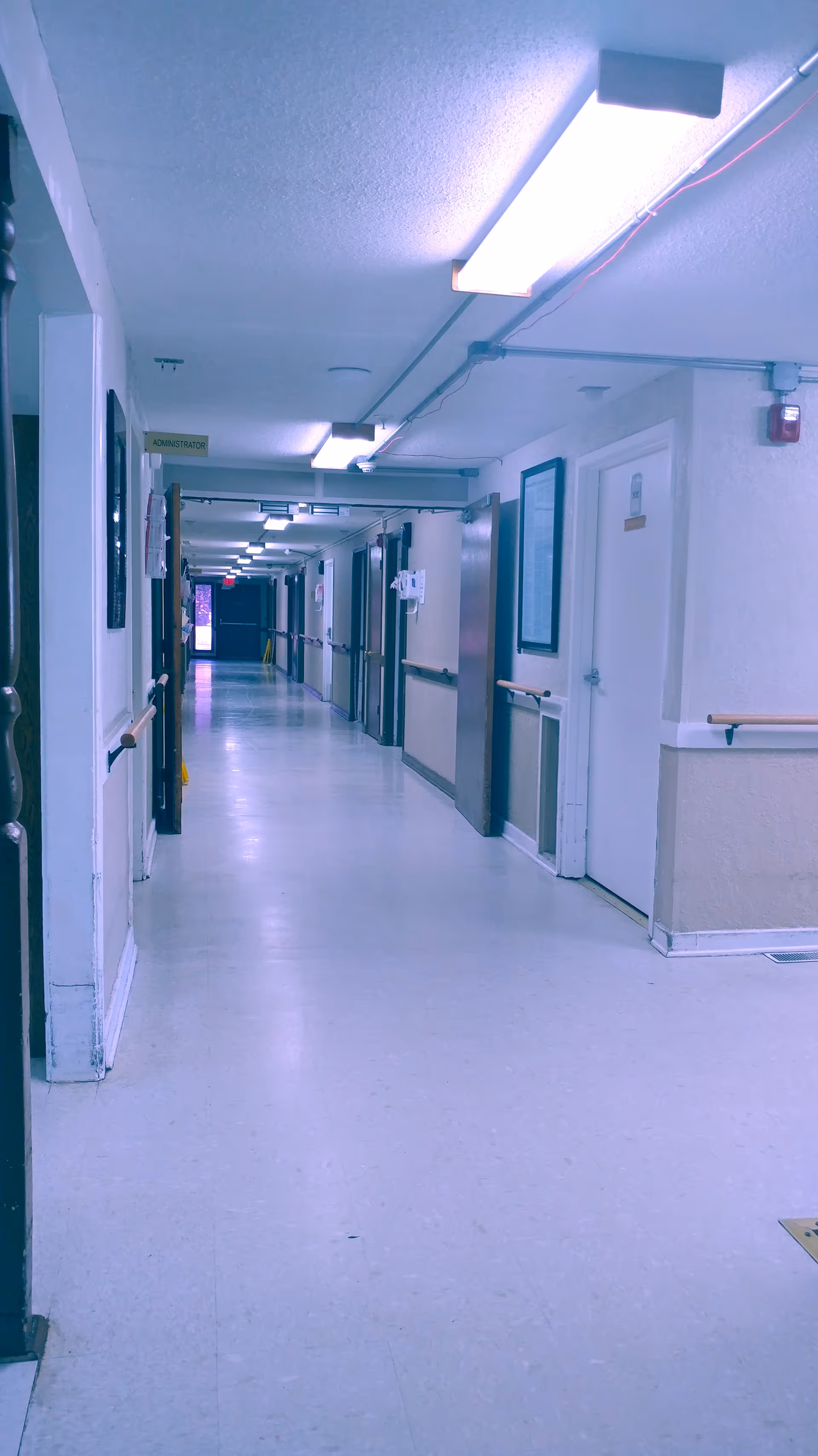 A long, empty hallway in a healthcare facility with white walls, handrails on both sides, several closed doors, overhead fluorescent lighting, and a sign that reads 'ADMINISTRATOR' on the left side.