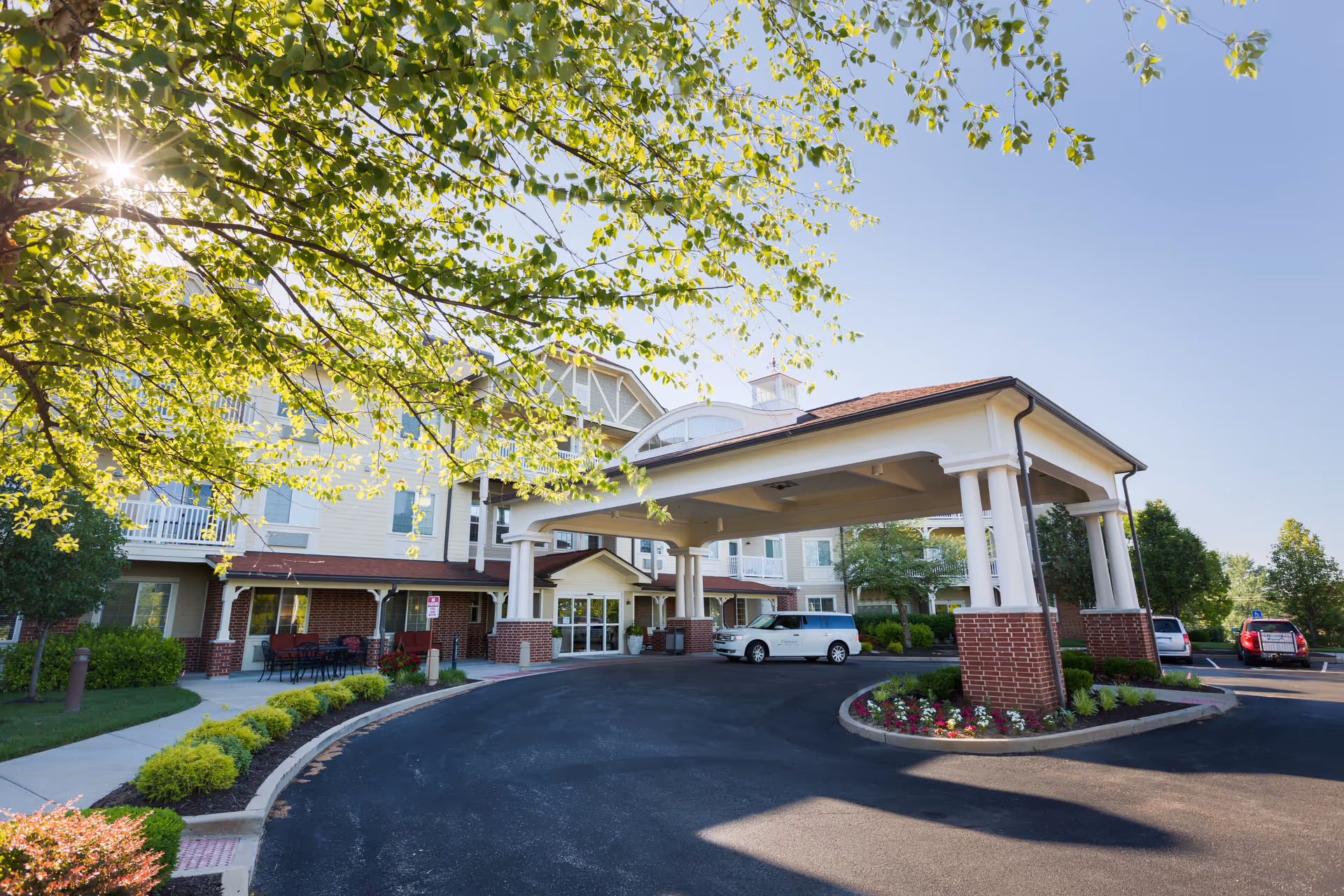 Entrance of Crestview Senior Living facility with a covered drop-off area supported by white columns and brick bases. The building is surrounded by well-maintained landscaping including bushes, flowers, and trees with sunlight filtering through the leaves. Several vehicles are parked near the entrance under a clear blue sky.