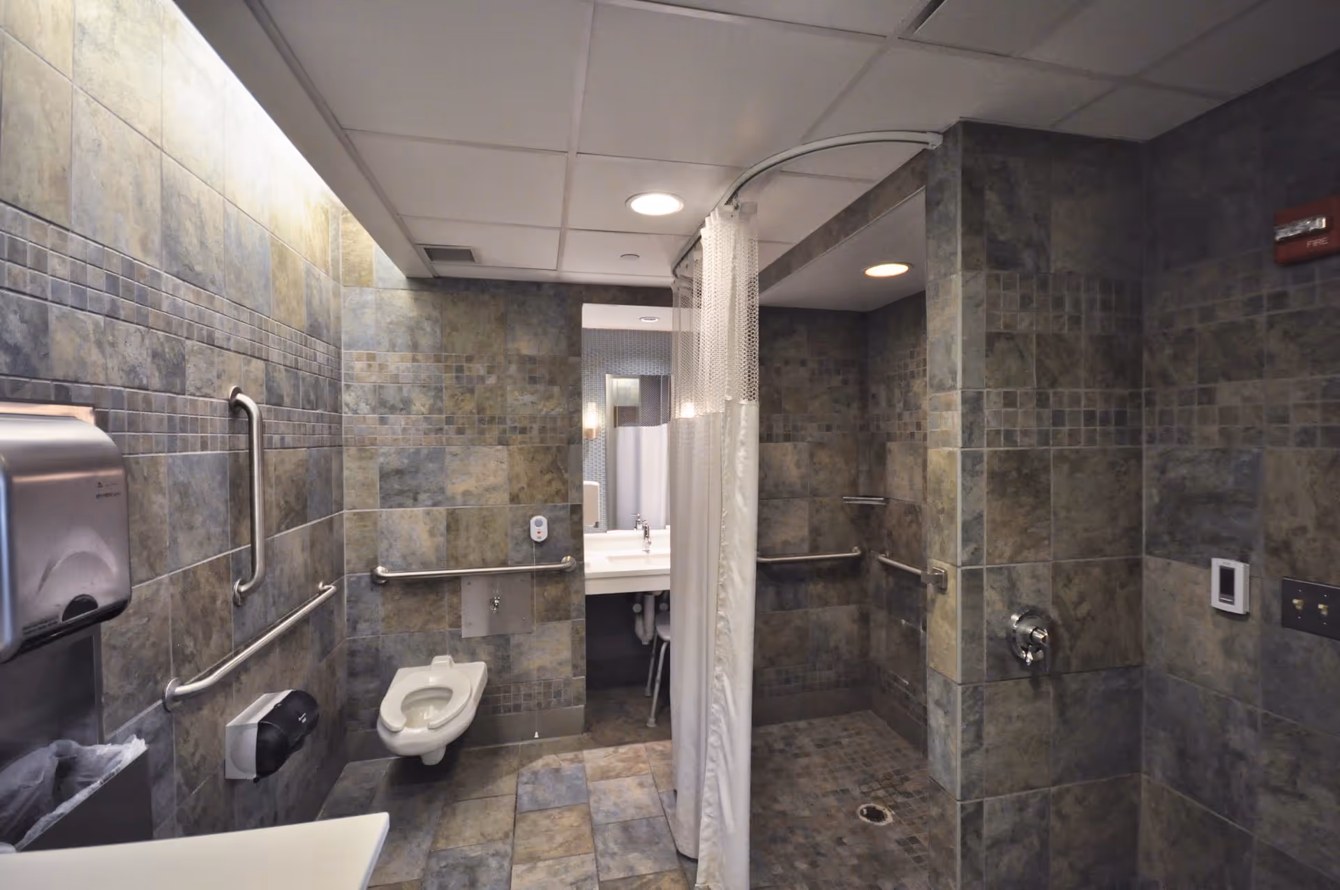 A tiled bathroom with a wall-mounted toilet, grab bars, a paper towel dispenser, a soap dispenser, a sink with a mirror, and a shower area separated by a white curtain. The walls and floor are covered in gray and beige tiles.