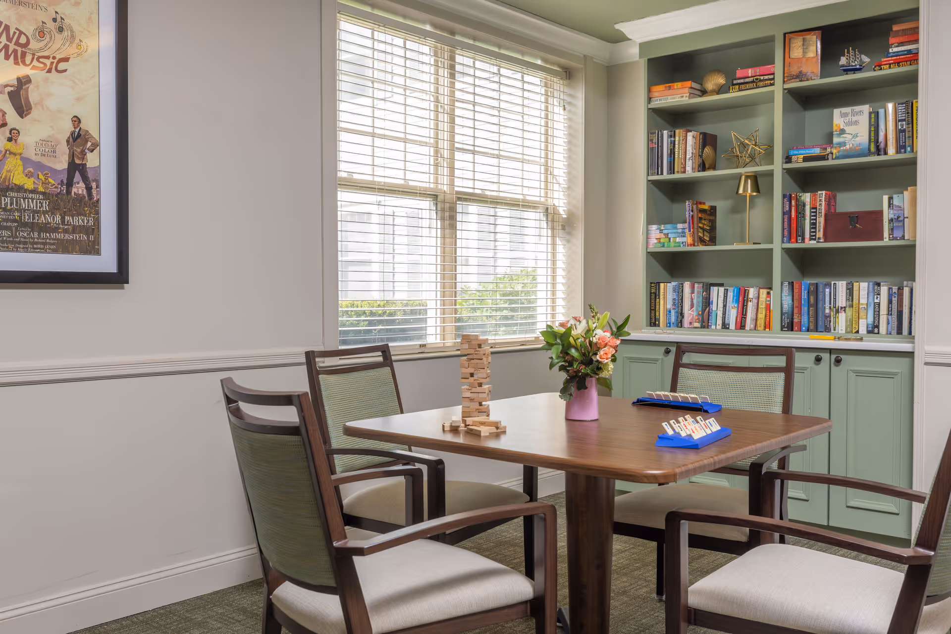 A sunlit communal room with a wooden table and chairs, built-in bookshelves, a window with blinds, and tabletop games and flowers.