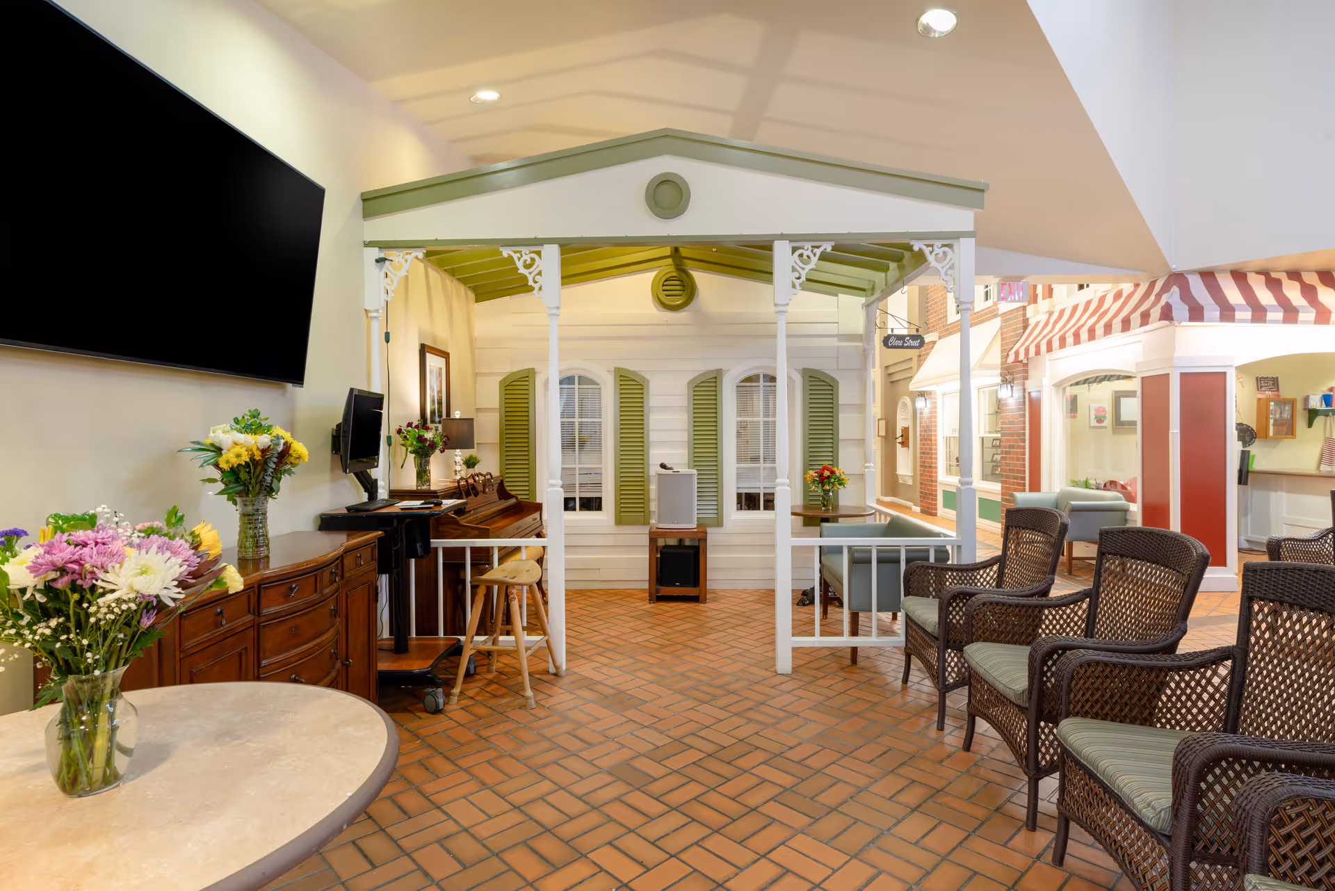 Interior common area of a senior living facility featuring a cozy seating arrangement with wicker chairs and cushioned seats, a small table with a vase of flowers, a large flat-screen TV mounted on the wall, and a decorative structure resembling a small house with green shutters and a green roof. The floor is tiled with reddish-brown bricks, and there are additional seating areas and decor visible in the background.