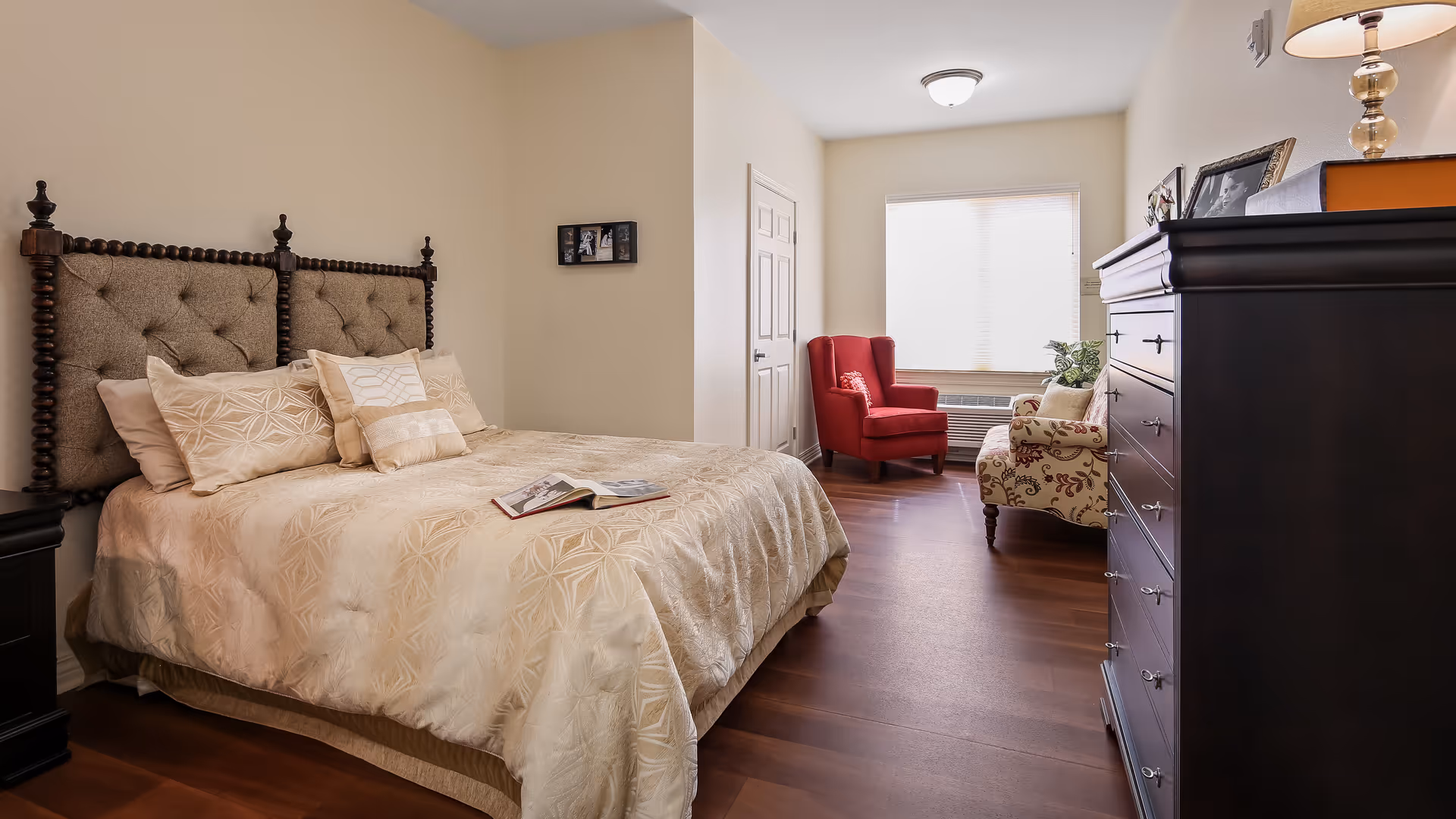 A cozy bedroom with a large bed featuring a tufted headboard and beige patterned bedding. There are several pillows on the bed and an open book resting on the bedspread. To the right, there is a dark wooden dresser with framed photos and a lamp on top. In the background, a red armchair and a floral upholstered chair are positioned near a window with blinds. The room has wooden flooring and neutral-colored walls.