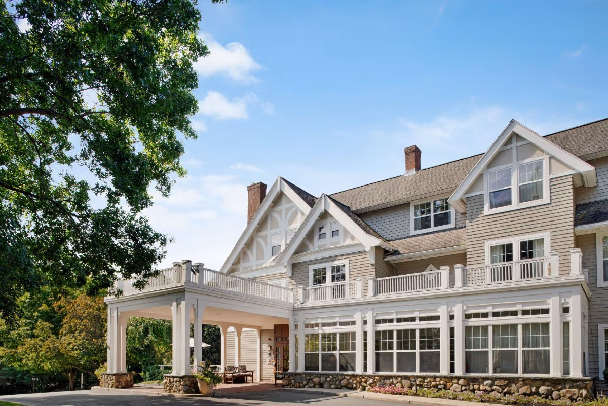 Exterior view of a large, multi-story residential building with beige siding, white trim, and a covered entrance supported by white columns with stone bases. The building is surrounded by trees and greenery under a blue sky with some clouds.