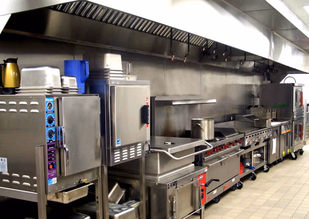 Commercial stainless-steel kitchen with ovens, stovetops, large ventilation hood, and cooking equipment lined up along a tiled floor.