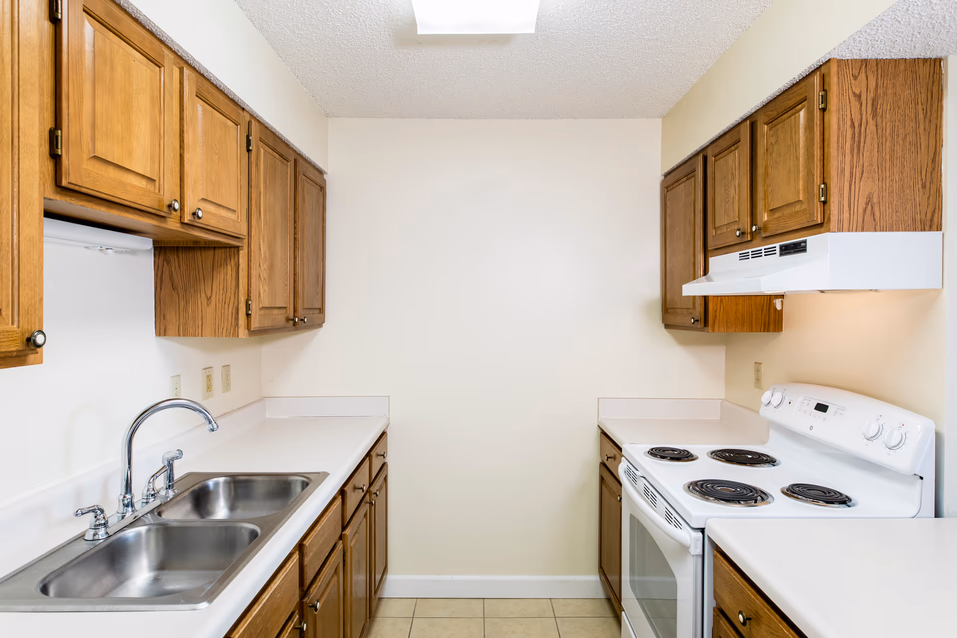 A small kitchen with wooden cabinets, white countertops, a double stainless steel sink with a faucet on the left side, and a white electric stove with four burners and an oven on the right side. The walls are light-colored, and the floor is tiled. There is a white range hood above the stove.