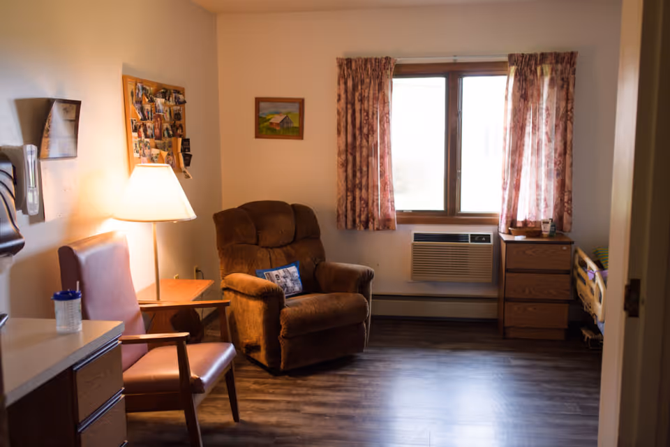 Small senior living room with a recliner, armchair and side table beside a window with floral curtains and a dresser.