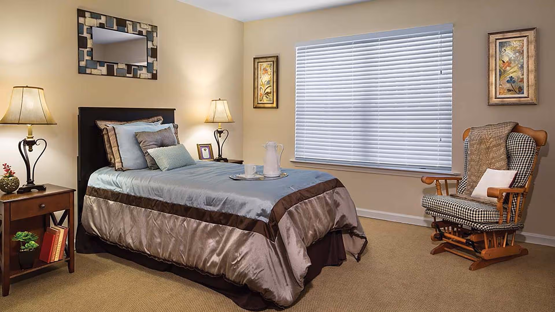 A neatly made single bed with multiple pillows and a tray with a pitcher and cup on it in a cozy bedroom. The room features two bedside tables with lamps, a cushioned rocking chair with a throw blanket and pillow, a large window with closed blinds, and framed artwork on the walls.