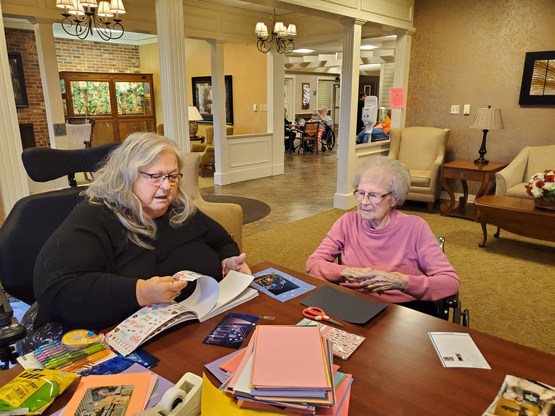 Two elderly women sitting at a table in a well-lit common area of a senior living facility. One woman with long gray hair and glasses is looking through a book or magazine, while the other woman with short gray hair and glasses is sitting in a wheelchair, watching. The table is covered with colorful paper, scissors, and craft supplies. The background shows a spacious room with chairs, tables, and other residents.