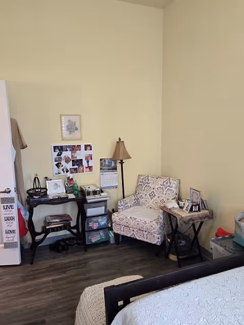 A cozy corner of a bedroom featuring a patterned armchair next to a small side table with framed photos and a basket underneath. A floor lamp stands behind the chair. To the left, there is a small black table with various items including a calendar, photos pinned on a board, and storage drawers underneath. The room has light yellow walls and wood flooring, with part of a bed visible in the foreground.