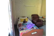 Small nursing-home bedroom with a single bed covered in colorful blankets and a stuffed animal, a recliner chair, and floral wallpaper.