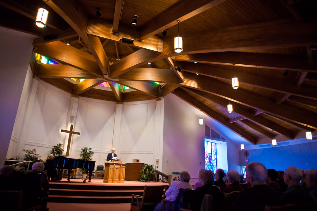 Interior of a chapel with wooden beams on the ceiling, a large cross on the wall, stained glass windows, and an elderly man standing at a wooden podium addressing an audience seated in chairs.