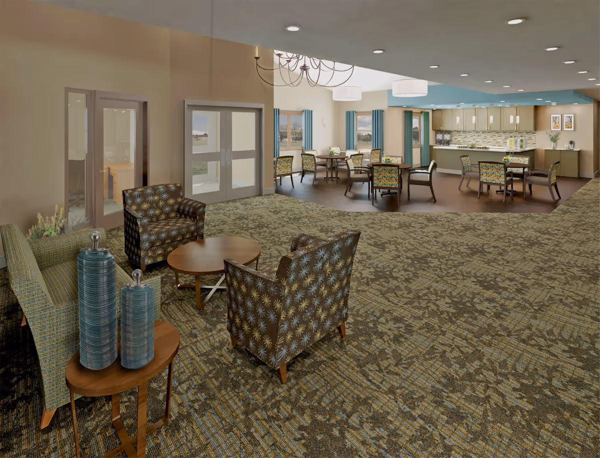 A spacious senior living facility common area featuring a seating area with patterned armchairs and a sofa around a round wooden coffee table. In the background, there is a dining area with several tables and chairs, large windows with blue curtains, and a kitchen area with cabinets and a countertop. The room has a patterned carpet and neutral-colored walls with recessed lighting and ceiling fixtures.