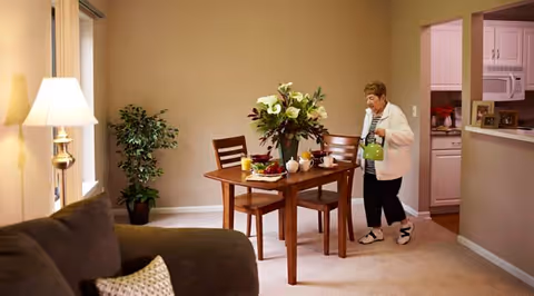A senior woman pouring tea from a green teapot into a cup at a wooden dining table set with breakfast items and a floral centerpiece in a cozy living and dining area. There is a brown couch with a patterned pillow, a floor lamp, and a potted plant near a window. The kitchen is visible through an open doorway.