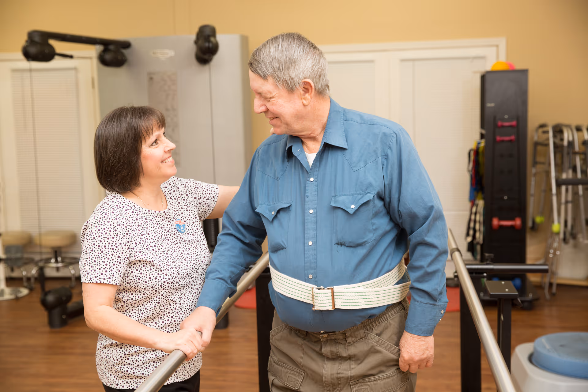 A female caregiver assists an elderly man with physical therapy in a rehabilitation room. The man is wearing a blue shirt and a gait belt while holding onto parallel bars. The caregiver is smiling and supporting him as he practices walking.