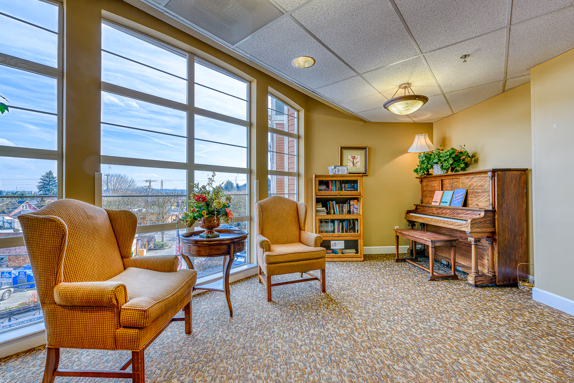 A cozy senior living common area with large windows letting in natural light. The room features two upholstered armchairs and a small round wooden table with a floral arrangement. Against the wall is an upright wooden piano with sheet music on the stand, a wooden bench, a bookshelf filled with books, a table lamp, and a potted plant on top of the piano. The carpet is patterned and the walls are painted a warm beige color.