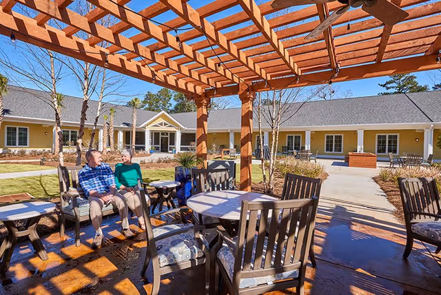 Outdoor seating area at a senior living facility with wooden pergola overhead, several cushioned chairs and tables, and two elderly people sitting and talking. The background shows a single-story building with multiple windows and a clear blue sky.