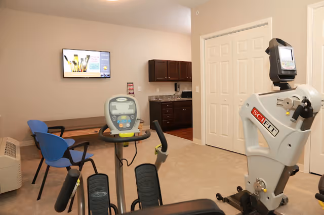 A small exercise room with fitness equipment including a stationary bike and a SciFit machine. There are two blue chairs and a wooden bench against the wall. A flat-screen TV is mounted on the beige wall, and a kitchenette with dark cabinets and a granite countertop is visible in the background.