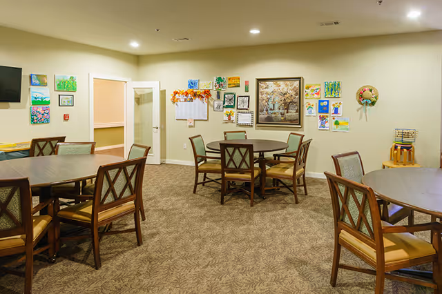 A well-lit room with multiple round wooden tables surrounded by cushioned chairs. The walls are decorated with various colorful paintings and artwork, including a large framed picture and a wreath. There is a door with a glass panel and a small TV mounted on the wall.