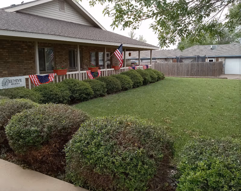 Single-story brick senior living facility front with a covered porch, shrubs, and flags.