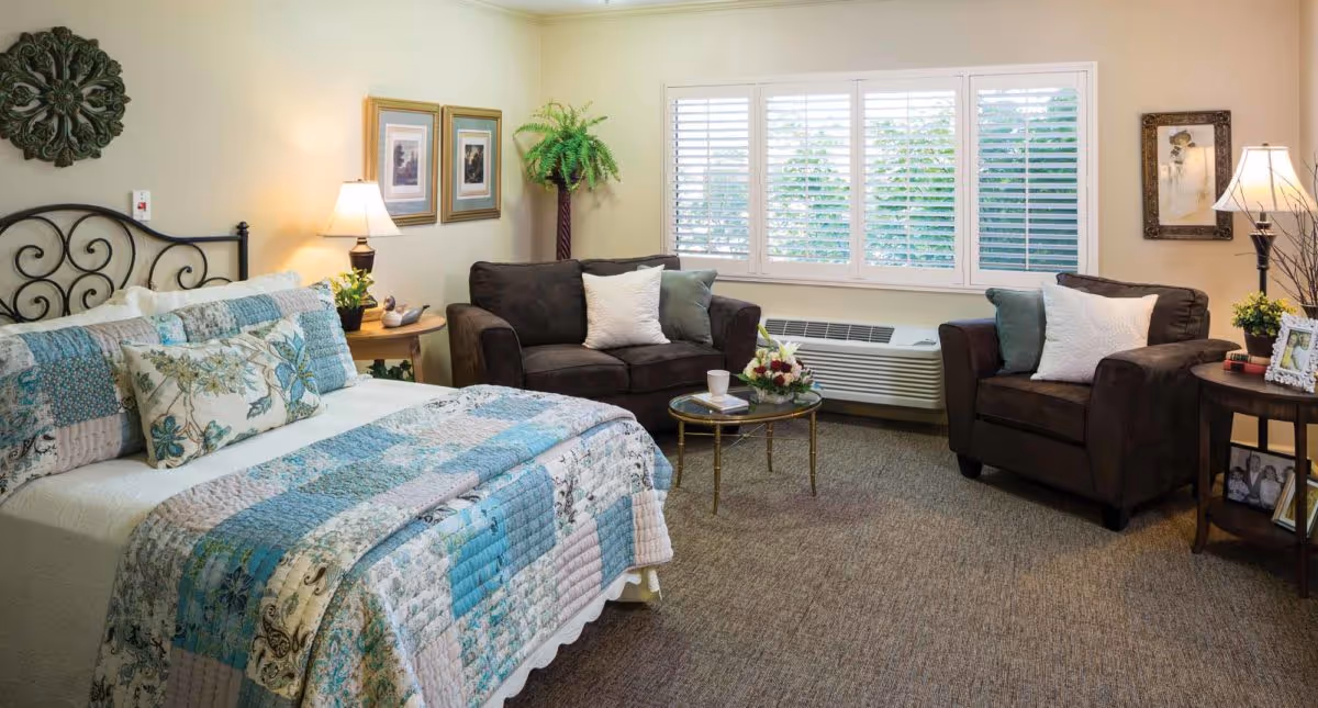 A cozy bedroom in Delaware Plaza Assisted Living Community featuring a bed with a blue and white patchwork quilt, two brown armchairs with white and green pillows, a small round glass coffee table with a flower arrangement and a cup, two side tables with lamps and framed photos, framed artwork on the walls, a large window with white shutters, and a potted plant in the corner.