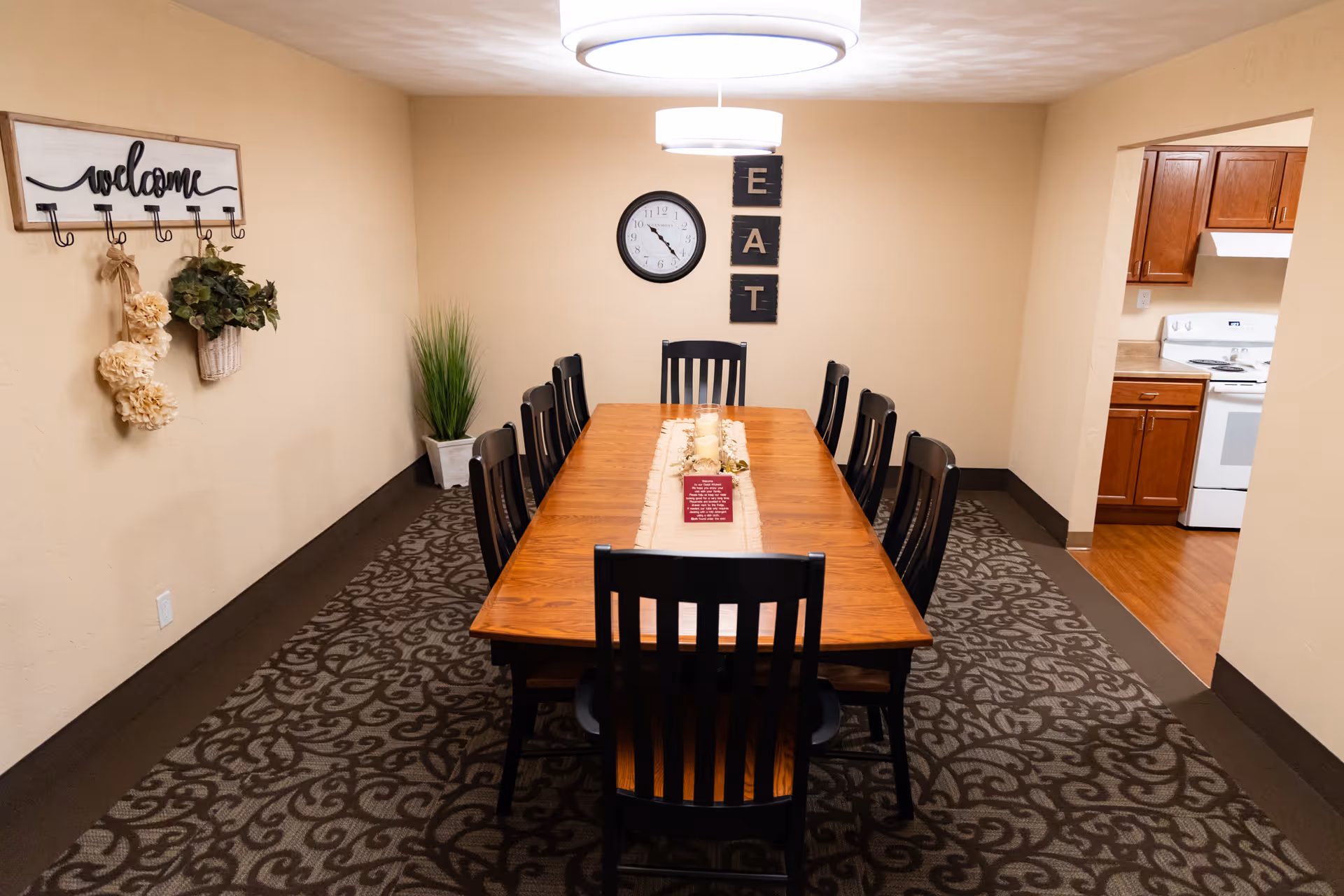 A dining room with a long wooden table surrounded by eight black chairs. The table has a beige runner with candles and a small decorative sign. On the wall at the far end, there is a clock and three black plaques spelling out the word 'EAT'. To the left wall, there is a decorative 'welcome' sign with hooks holding dried flowers and a small potted plant. The room has beige walls and patterned carpet. A kitchen with wooden cabinets and white appliances is visible through an open doorway on the right.