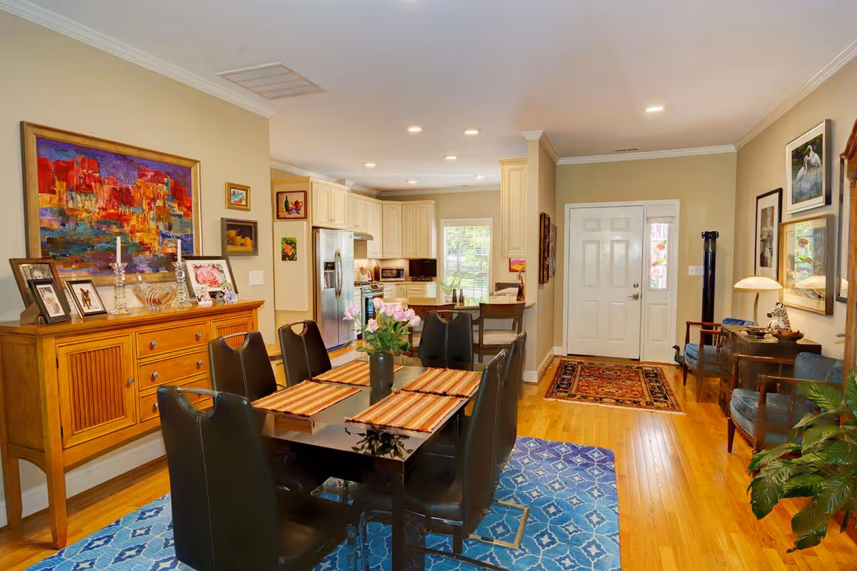 Open dining area with a dark table and six black chairs on a blue rug, a wooden sideboard with framed art, and a view into the kitchen and entryway.