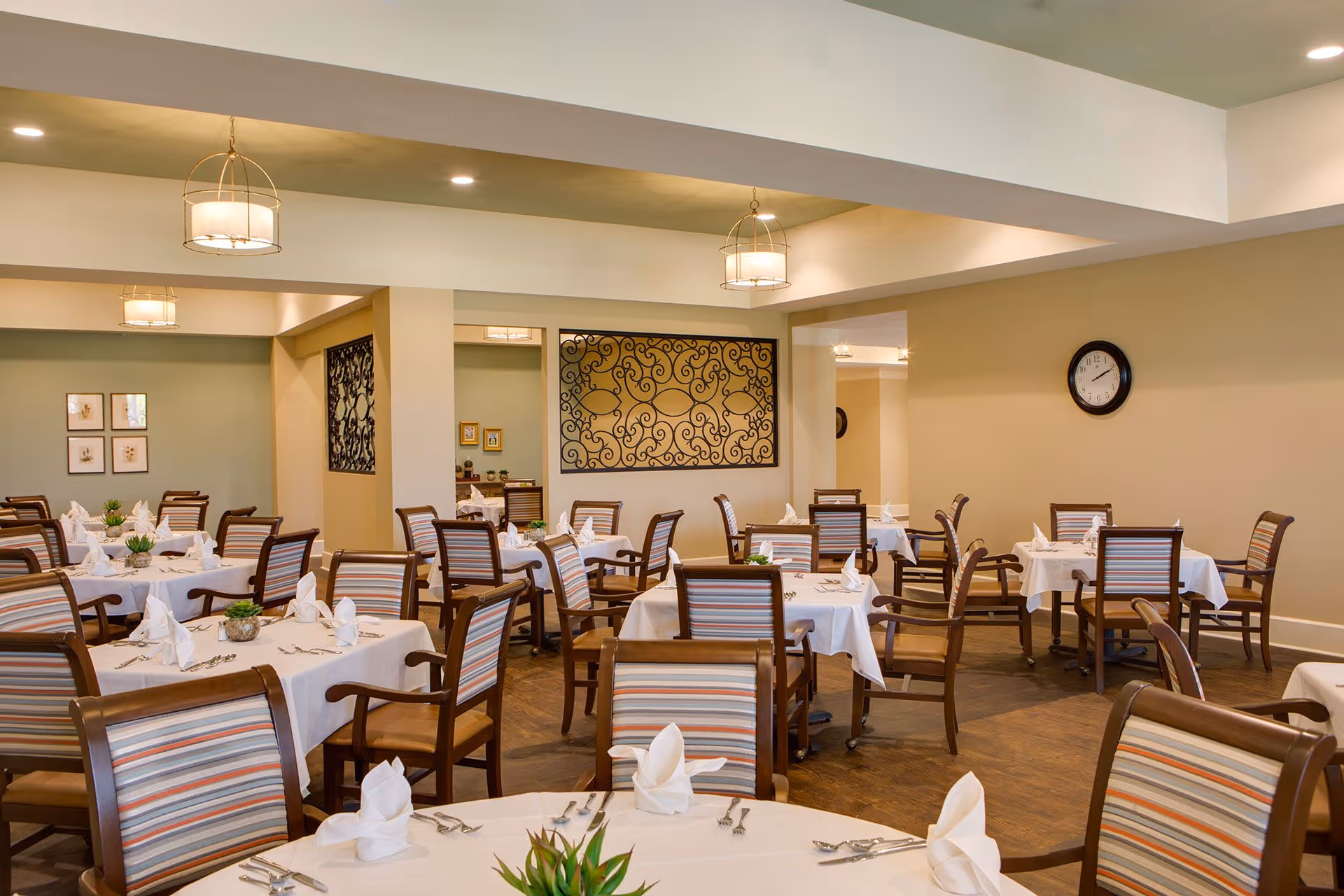 A spacious dining room with multiple tables covered in white tablecloths, each set with folded napkins, silverware, and small green plants as centerpieces. The chairs have wooden frames with striped upholstery. The room has beige walls, decorative wall art, a clock, and modern hanging light fixtures.